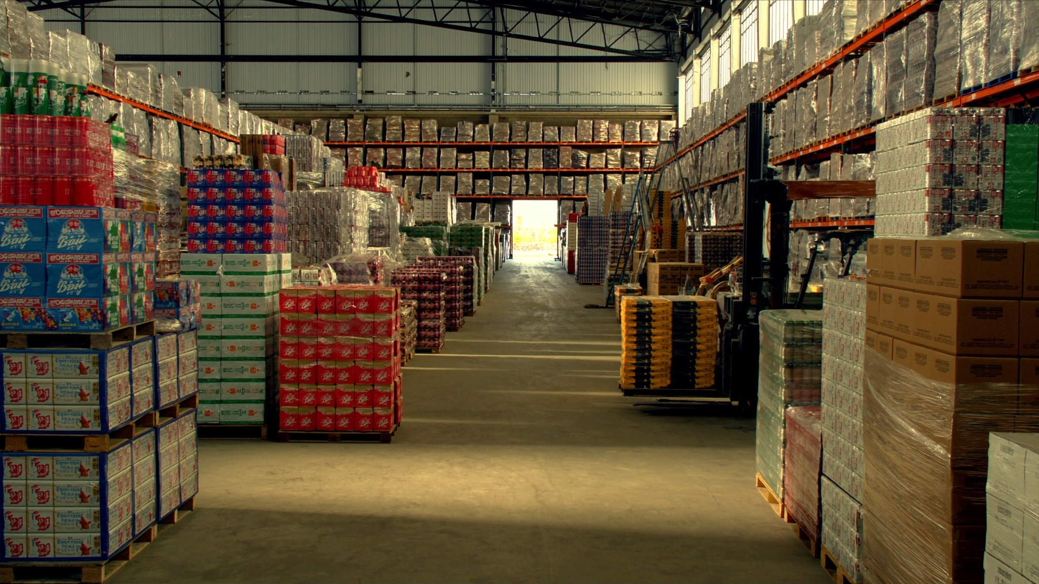 Wide editorial view of a modern American beverage distribution warehouse — pallets of beer and wine cases on industrial racking with natural daylight, illustrating the three-tier alcohol distribution system